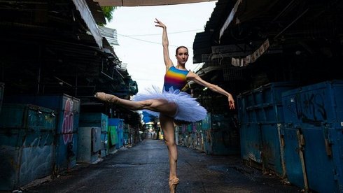 Na środku ciemnej, opuszczonej alejki stoi tancerka w kolorowym stroju w pozie baletnicy.