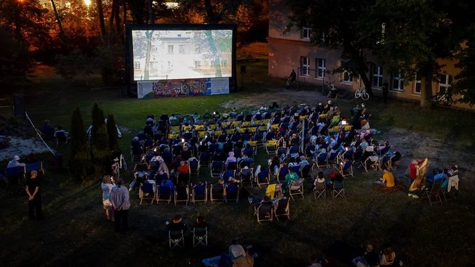 - Kadr wypełniają leżaki z widzami na łące. Osoby oglądają film wyświetlany na dużym ekranie. Wieczór jest wczesny. Mrok jeszcze nie zapadł.