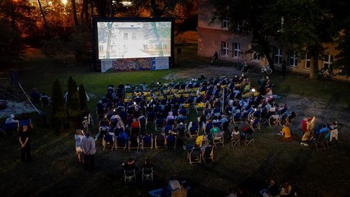 Kadr wypełniają leżaki z widzami na łące. Osoby oglądają film wyświetlany na dużym ekranie. Wieczór jest wczesny. Mrok jeszcze nie zapadł.