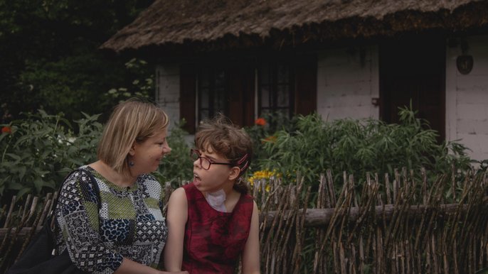 - Julia i pani Monika siedzą na ławce. W tle drewniany dom z ogrodem.