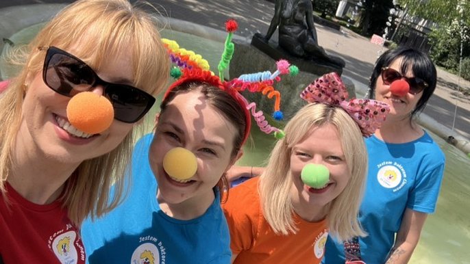 - Zdjęcie przedstawia cztery osoby w kolorowych koszulkach i z kolorowymi kulkami na nosach. Zdjęcie zrobione na tle fontanny i rzeźby siedzącej kobiety. W oddali zieleń, drzea i budynki.