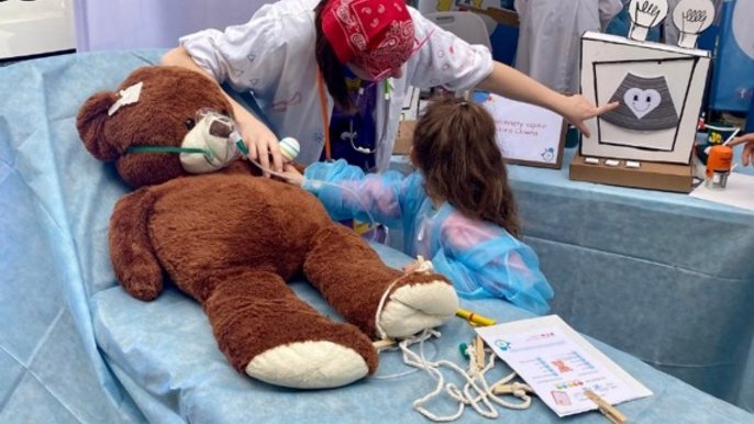 - Na zdjęciu widać stoisko Fundacji Dr Clown, organizacji znanej z działań na rzecz uśmiechu i wsparcia emocjonalnego dzieci w szpitalach. Na pierwszym planie znajduje się duży pluszowy miś leżący na niebieskim łóżku szpitalnym – wygląda, jakby był „pacjentem”. Obok niego stoi dziewczynka w niebieskim fartuchu ochronnym, która „bada” misia razem z dorosłą osobą w białym fartuchu, z czerwoną chustką na głowie i kolorowymi elementami stroju. W tle widać banery z logo Fundacji Dr Clown i hasłem „#uśmiechleczy