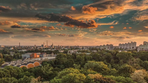 Panorama Łodzi na tle zachodzącego słońca. Ponad parkiem wysokie zabudowania na linii horyzontu.