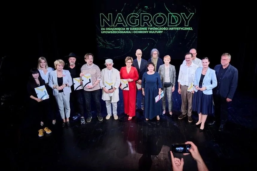 Na scenie teatralnej stoi grupa ludzi, którzy w rękach trzymają dyplomy. Są to laureaci nagród w 2023 roku. W tle napis: Nagrody za osiągnięcia w dziedzinie twórczości artystycznej, upowszechnia i ochrony kultury.