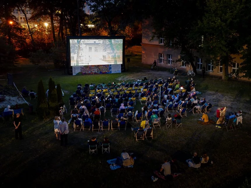 Kadr wypełniają leżaki z widzami na łące. Osoby oglądają film wyświetlany na dużym ekranie. Wieczór jest wczesny. Mrok jeszcze nie zapadł.